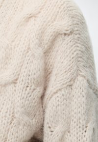 Gros plan d'un tissu tricoté épais et doux de couleur crème, avec un motif torsadé texturé et des fibres duveteuses.