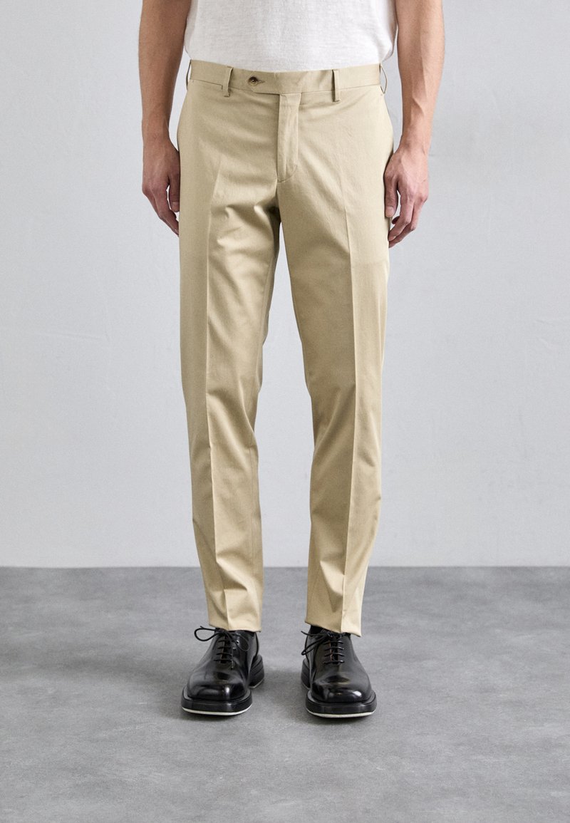 Béžové chino kalhoty s rovně střiženým designem, zapínání na knoflík a boční kapsy. Nosené s černými koženými botami. Hladká textura.