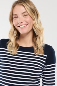 Lächelnde Frau mit blonden, welligen Haaren, die ein navy- und weißgestreiftes Langarmshirt trägt, vor einem einfarbigen Hintergrund.