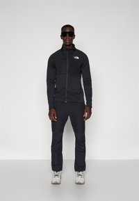 Melnā rāvējslēdzēja jaka un saskaņotas bikses no teksturēta auduma, ar priekšējo kabatu un The North Face logotipu. Valkā ar pelēkām un baltām sporta apaviem.
