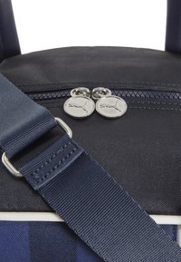 Nærbillede af en sort og blå stoftaske med en justerbar navy strop og sølvfarvede lynlås-dragere med en puma-logo.