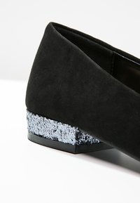 Chaussure noire en daim à talon bas avec un talon bloc couvert de paillettes argentées sur fond blanc.