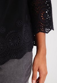 Blusa nera con dettagli in pizzo intricati e orlo a scallop; presenta motivi floreali e tessuto trasparente sulle maniche. Visibili pantaloni grigi.