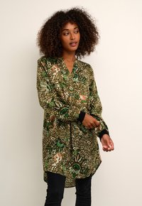 Chemise longue à motif vert avec un décolleté en V, des motifs floraux et animaliers, et des poignets noirs. Fabriquée dans un tissu lisse avec une coupe ample.