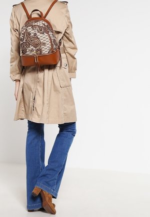 Personne portant un trench-coat beige, un jean bleu, des bottes à talons brunes et un petit sac à dos brun à motif, se tenant devant un fond blanc.