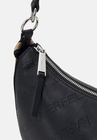 Angolo di una borsa in pelle nera con cerniera argentata, targhetta con logo perforato e anello metallico che attacca una tracolla in tessuto a strisce.