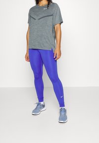 Maglietta sportiva grigia con texture e maniche corte, abbinata a leggings blu aderenti e scarpe da corsa grigie con accenti bianchi.