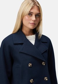 Navy wool coat with a double-breasted design, large lapels, and brass buttons. Smooth texture, featuring side pockets and a tailored fit.