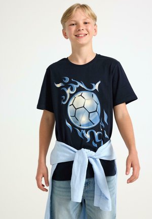 Marineblå T-shirt med et fodboldmotiv og flamme designs i lyseblå nuancer. Bæres med en lyseblå skjorte bundet om livet.