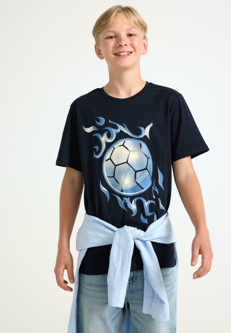 Marineblå T-shirt med et fodboldmotiv og flamme designs i lyseblå nuancer. Bæres med en lyseblå skjorte bundet om livet.