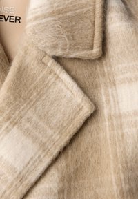 Cappotto di lana beige caratterizzato da una morbida texture, un ampio colletto e un sottile motivo a quadri. Cuciture visibili e rever sartoriali.