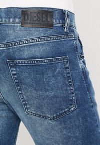 Vaqueros de mezclilla azul con un acabado texturizado, dos bolsillos traseros y un parche de logo de cuero negro en la cinturilla.