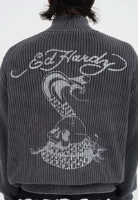 Person, der einen grauen Strickpullover mit einem weißen gestickten Kobra- und Schädeldesign sowie kursivem "Ed Hardy"-Text auf dem Rücken trägt.