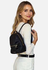 Zaino in nylon nero dalla forma arrotondata, con dettagli in zip dorate e una tasca frontale. Indossato su una spalla, abbinato a un maglione beige.