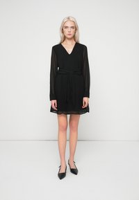 Γυναίκα με μακριά ξανθά μαλλιά, ντυμένη με μαύρο φόρεμα με V-πλευρά και μακριά μανίκια, με ζώνη που δένει στη μέση και μαύρες μυτερές γυναικείες γόβες, στέκεται σε λευκό πάτωμα.