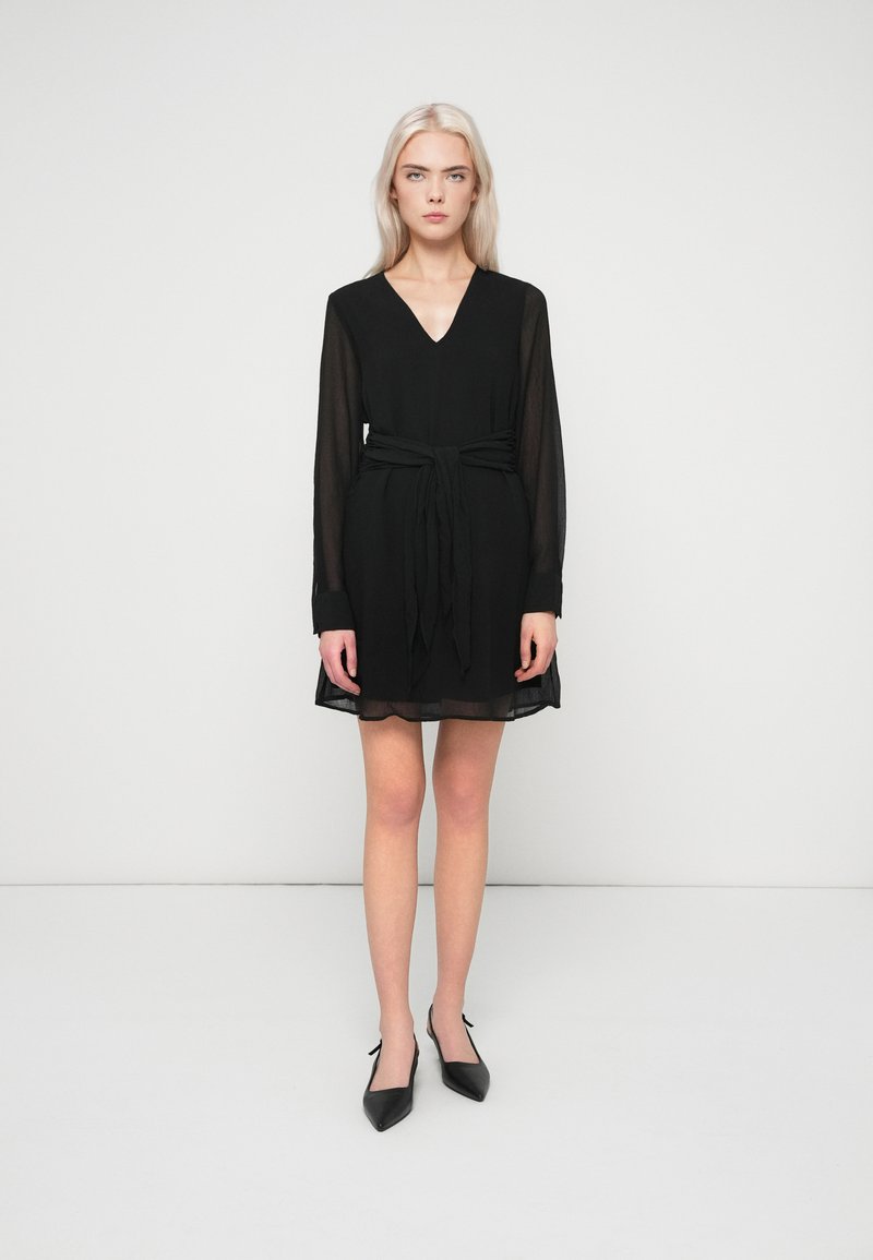 Γυναίκα με μακριά ξανθά μαλλιά, ντυμένη με μαύρο φόρεμα με V-πλευρά και μακριά μανίκια, με ζώνη που δένει στη μέση και μαύρες μυτερές γυναικείες γόβες, στέκεται σε λευκό πάτωμα.