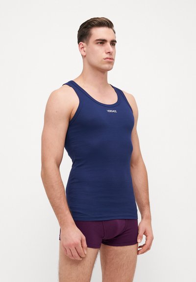 Navyblaue Tanktop mit breiten Trägern und enganliegendem Design, hergestellt aus einem glatten Stoff. Mit einem kleinen Logo auf der Brust. Kombiniert mit lila Unterwäsche.