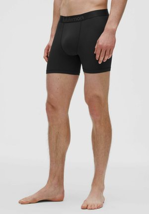 Uomo in piedi a piedi nudi che indossa boxer neri con fascia in vita con marchio su uno sfondo chiaro e uniforme.