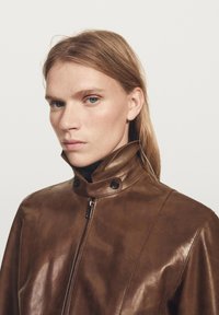 Brown leather jacket with a high collar, zip closure, and two snap buttons at the neck. Smooth texture with subtle sheen.