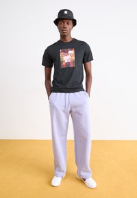 Schwarzes T-Shirt mit einem zentralen Grafikdesign, kombiniert mit helllila Jogginghosen. Das Model trägt einen schwarzen Bucket Hat und weiße Sneakers.