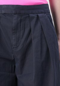 Dunkelblaue, hoch taillierte Hose mit Falten, mit glatter Textur und seitlichen Taschen, kombiniert mit einem strukturierten rosa Oberteil.