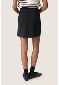 Zwarte mini-jurk met een gladde afwerking, met een ontspannen tailleband. Het model draagt zwarte sokken en een zacht, geplooid donker schoeisel.