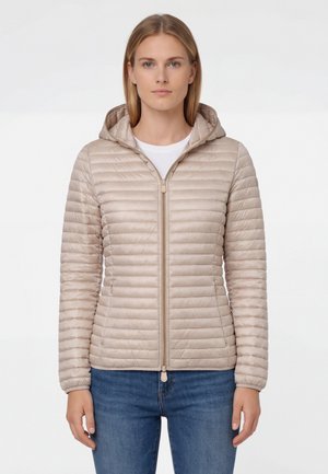 Vrouw draagt een beige doorgestikte jas met capuchon, dichtgeritst, gecombineerd met een blauwe spijkerbroek, staand tegen een effen witte achtergrond.