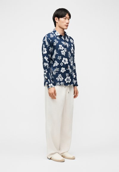 Άνδρας που στέκεται φορώντας μπλε σκούρο πουκάμισο με μακριά μανίκια, με λευκά λουλουδάτα και ζωικά σχέδια, άσπρο φαρδύ παντελόνι και ανοιχτόχρωμα παπούτσια slip-on.