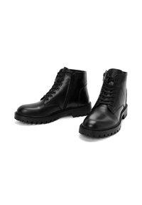Par de botas de cuero negras hasta el tobillo con cordones y cremalleras laterales, que cuentan con suelas de goma gruesas y con relieve sobre un fondo blanco.