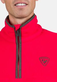 Red fleece pullover with high collar, grey zipper, and black logo patch. Soft texture, featuring a half-zip design for easy wear.
