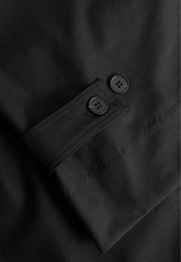 Melnā audumā ir pogāšanai paredzēts aproce ar divām melnām pogām, gluda tekstūra un izšūti akcenti gar malu.