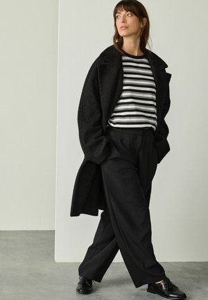 Femme portant un pantalon large noir, des chaussures noires, un haut rayé noir et blanc, et un long manteau noir, se tenant sur un sol en béton.