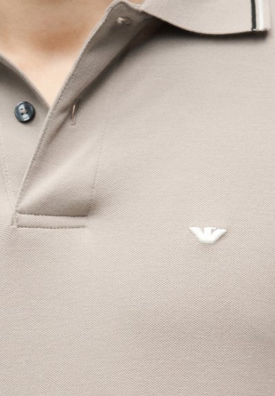 Μπεζ πόλο πουκάμισο με μαύρο κουμπί και μικρό λευκό κεντημένο λογότυπο στο στήθος, εμφανιζόμενο στο επάνω μέρος του κορμού ενός ατόμου.
