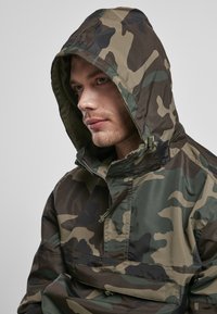 Camouflage windbreaker jas in tinten van groen en bruin, met een capuchon, halve rits aan de voorkant en een textuur binnenin.