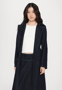 Mørk denim trenchcoat med grønn søm, dobbeltspent design og store knapper. Båret over en hvit, kort genser.