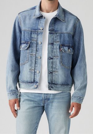 Mand iført en lyseblå denimjakke med knapklappede lommer over en hvid t-shirt og lyseblå jeans.