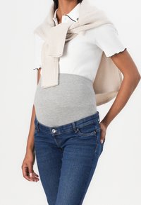 Těhotenský outfit s bílým polokošilí, šedým elastickým pásem a tmavě modrými džíny. Světle béžový pletený svetr je přehozený přes ramena.