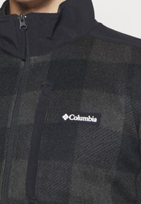 Veste polaire Columbia à carreaux noirs et gris avec une fermeture éclair frontale et une poche poitrine zippée, portée par une personne.