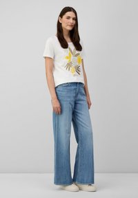 T-shirt blanc avec un motif floral comprenant des accents jaunes, associé à un jean large bleu clair et des chaussures à enfiler beiges.