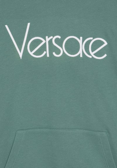 Grön sweatshirt med en stor vit "Versace"-logotyp. Materialet är textilerat, med en framficka och ribbad detalj vid nederkanten.