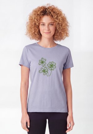 Femme aux cheveux bouclés portant un t-shirt violet clair avec trois illustrations vertes de trèfles à quatre feuilles sur le devant.