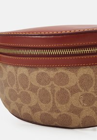 Borsa in tessuto marrone con motivo circolare testurizzato, caratterizzata da una parte superiore in pelle rossa scura e dettagli in zip dorata.