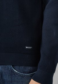 Granatowy sweter z fakturowanej tkaniny, z prążkowanymi mankietami i dolnym brzegiem. Posiada małą metkę z napisem "Elevated Casual" na dolnej krawędzi.