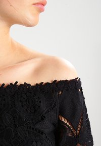 Haut en dentelle noire à épaules nues, mettant en avant des motifs floraux et des bords festonnés, mettant en valeur une texture complexe et une coupe ajustée.