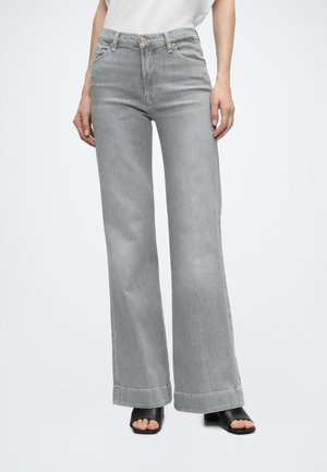Femme portant un jean gris taille haute à jambes larges avec ourlets retournés et des sandales noires à talons blocs ouvertes aux orteils, debout devant un fond uni.