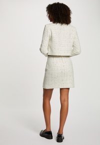 Outfit in tweed bianco con giacca corta e gonna aderente. Presenta un motivo testurizzato, design minimalista e zip sul retro. Scarpe nere.