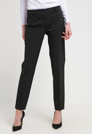 Femme portant un pantalon noir ajusté, une chemise blanche à manches longues et des chaussures à talons noirs, debout devant un fond uni.