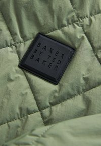 Rudens zaļas tonis audums ar matētu tekstūru, kurā ir taisnstūrveida melna etiķete ar uzrakstu "BAKER BY TED BAKER".