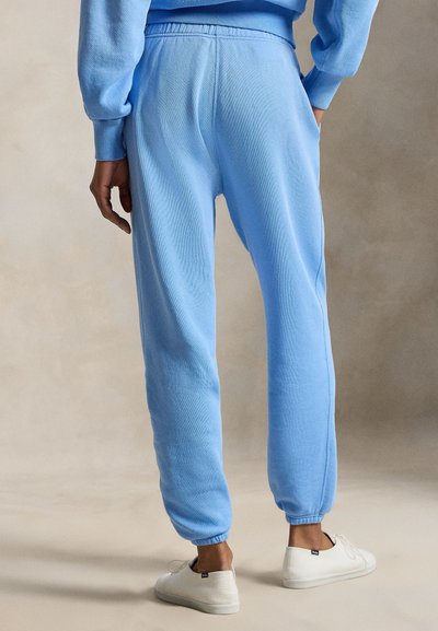 Άτομο που φοράει ανοιχτό μπλε φόρμα και αντίστοιχο φούτερ, στέκεται με τα χέρια στα πλάγια, άσπρα αθλητικά παπούτσια σε ουδέτερο φόντο.