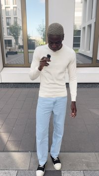 Pull en maille crème, jeans bleu clair, baskets noires et crème. Tenant un petit microphone noir. Debout sur une surface pavée.